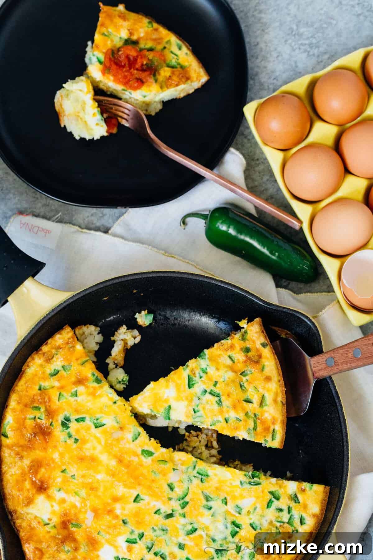This one-pan cheddar jalapeno tater tot frittata recipe puts a fun twist on a potato and egg frittata, baked inside a crispy tater tot crust. Overhead view of a slice of tater tot egg frittata next to the rest of a frittata in a cast iron skillet, next to another slice of frittata on a black plate and a carton of eggs.