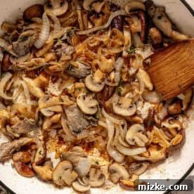 Velvety Mushroom Chicken 5 Sliced mushrooms and onions sautéing in a white pan with a wooden spatula, garnished with fresh herbs.
