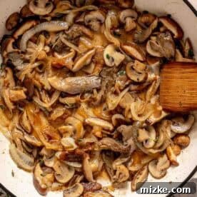 Velvety Mushroom Chicken 6 Sautéed mushrooms and onions in a white skillet with a wooden spatula.