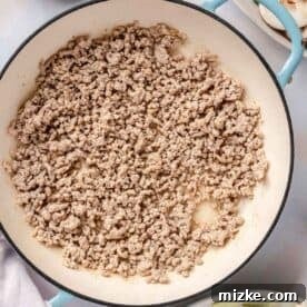 Ground pork being broken into smaller pieces and browned in an enameled cast iron skillet.