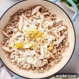 Garlic, onion, ginger, and sliced mushrooms added on top of the cooked ground pork in an enameled cast iron skillet.