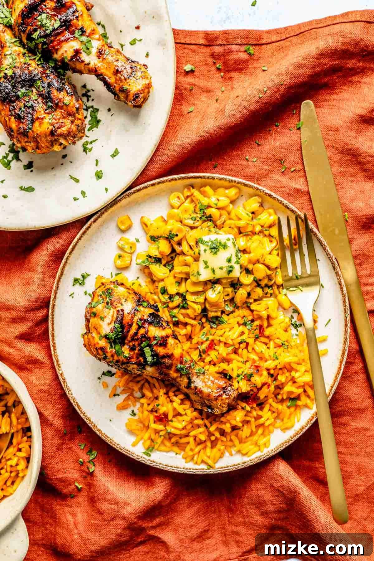 Zesty Peri Peri Chicken Drumsticks 3 Grilled chicken drumsticks and yellow rice with corn on a white plate, garnished with parsley, placed on an orange cloth. Gold fork and knife are beside the plate.
