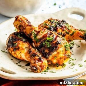 Grilled chicken drumsticks garnished with herbs on a white plate, with a side of corn and a bowl of rice in the background.