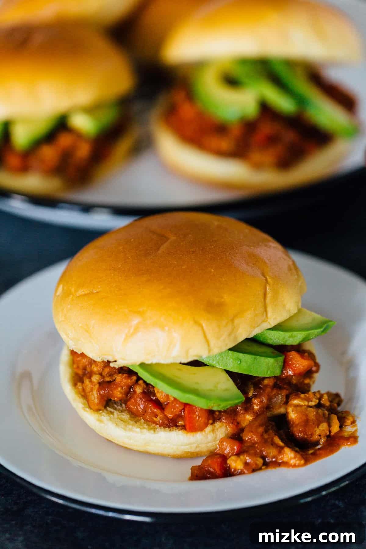 These pumpkin BBQ sloppy joes are going to be your new favorite weeknight recipe! They're a quick dinner idea filled with loads of flavor. A close-up shot of a pumpkin BBQ sloppy joes sandwich, garnished with avocado slices, on a plate, with a soft-focus background of more sandwiches.