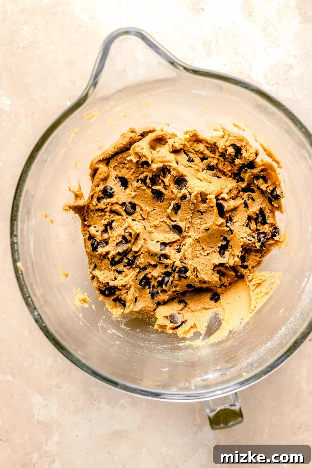 Caramelized Butter Chocolate Chip Cookies 7 Rich, glossy brown butter chocolate chip cookie dough in a glass mixing bowl, speckled with dark chocolate chips, ready for scooping.