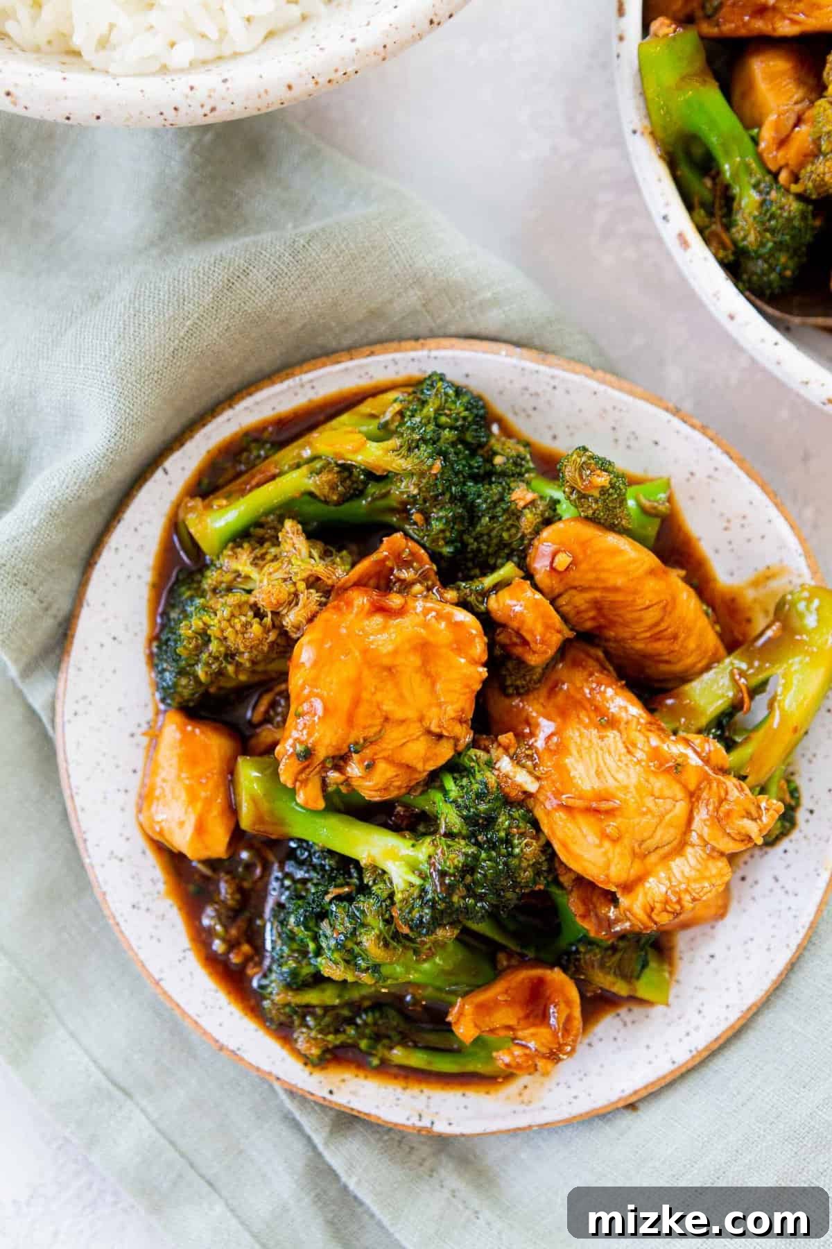A perfect serving of homemade Chinese Chicken and Broccoli, ready to enjoy with rice. Overhead shot of a single serving of Chinese Chicken and Broccoli on a small plate, accompanied by a portion of white rice, resting on a light green linen towel.
