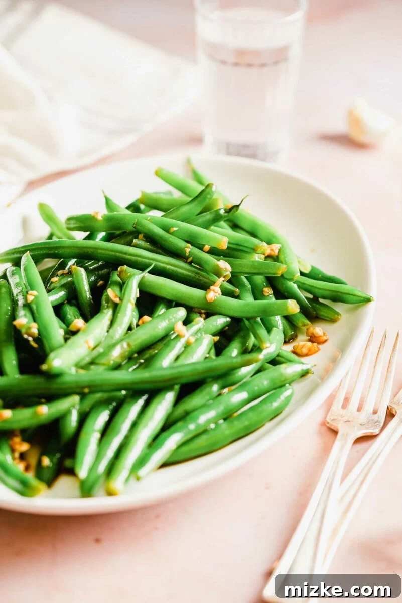 A fork and knife are artfully placed beside a generous serving of garlic and soy marinated green beans on a pristine white plate, inviting a taste.