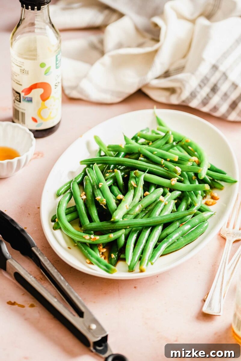 Tongs, a bottle of soy sauce, and elegant utensils are arranged next to a bowl of vibrant green beans, hinting at a delightful meal.