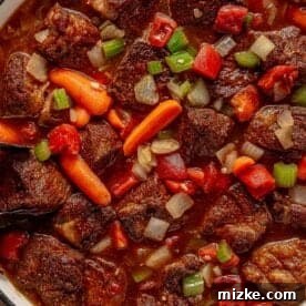 Hearty Ale Braised Beef 11 A close-up of a pot filled with beer braised pot roast. The stew contains chunks of beef, carrots, celery, onions, and diced tomatoes in a rich, brown broth. The colorful ingredients are mixed together, creating a hearty and appetizing appearance.