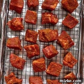 Hearty Ale Braised Beef 7 Seasoned pork belly cubes are evenly spaced on a wire rack, which is placed on a foil-lined baking sheet. The reddish-brown spice rub on the meat creates a rich, textured appearance.