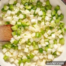 Hearty Ale Braised Beef 8 Close-up of a pot with diced onions, celery, and green bell peppers being stirred with a wooden spoon. The vegetables are uncooked, showing a mix of white and green colors. Carrot slices can be seen at the edge of the image.