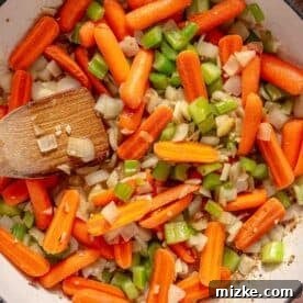 Hearty Ale Braised Beef 9 Overhead view of a pan filled with sautéed baby carrots, chopped celery, and onions. A wooden spatula is seen stirring the colorful vegetables.