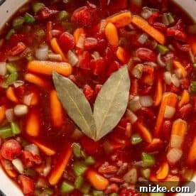 Hearty Ale Braised Beef 10 A close-up of a pot filled with colorful vegetable soup, featuring chopped tomatoes, carrots, celery, onions, and two bay leaves on top. The broth is rich and red, with various vegetables visibly submerged.