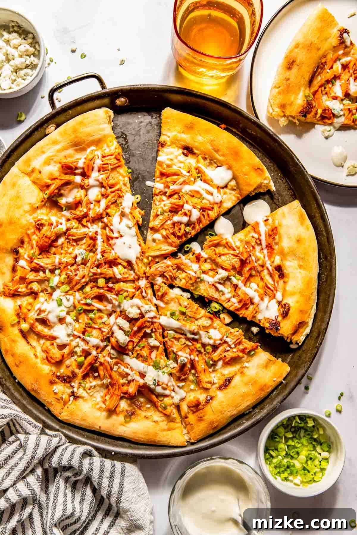 Kickin' Buffalo Chicken Pizza 2 A perfectly baked buffalo chicken pizza on a round pan, garnished with fresh green onions and a drizzle of white sauce. Accompanying it are a refreshing drink, a plate with a sliced serving, and buffalo sauce, all resting on a striped towel.