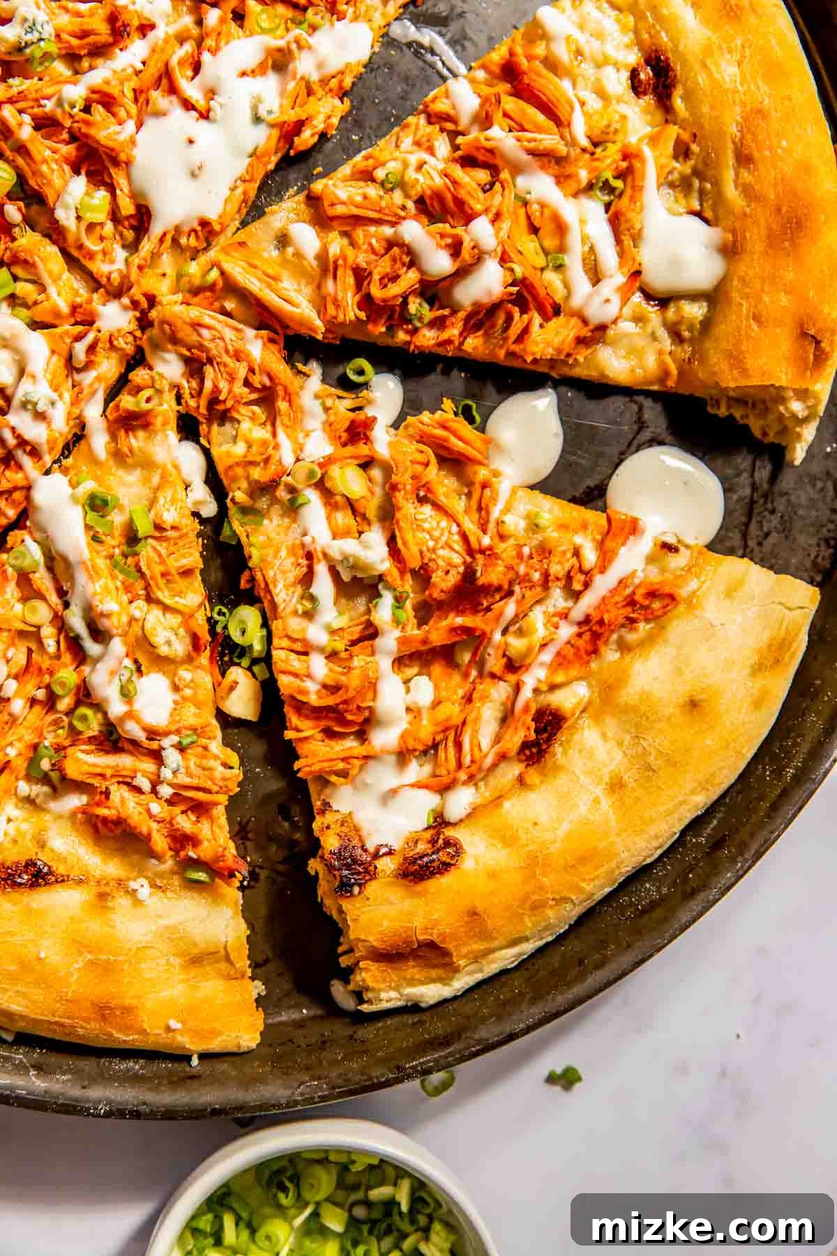 Kickin' Buffalo Chicken Pizza 3 A freshly baked pizza topped with shredded buffalo chicken, vibrant sliced green onions, and a delicate drizzle of white sauce, cut into perfect slices on a round baking pan.