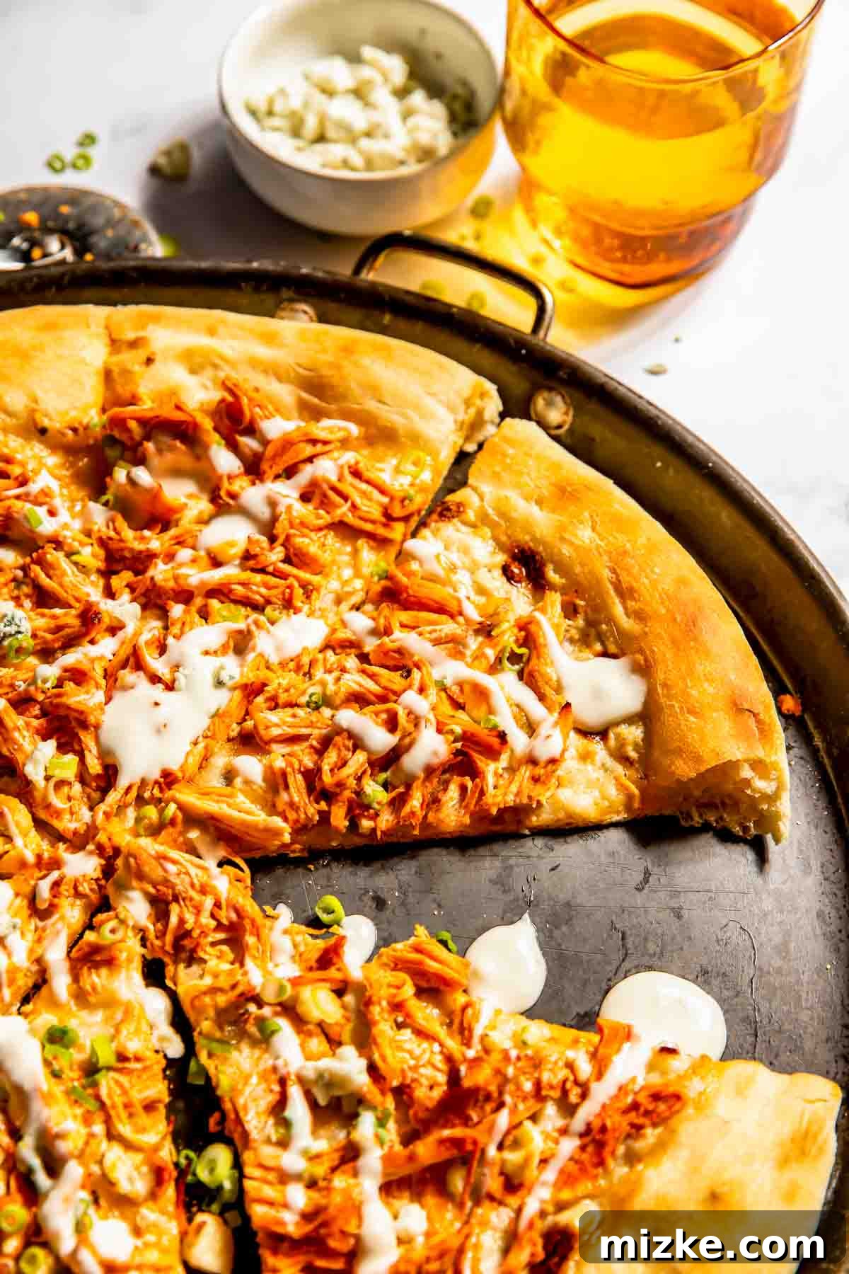 Kickin' Buffalo Chicken Pizza 4 A beautifully baked buffalo chicken pizza, artfully drizzled with white sauce and cut into neat slices, rests on a round baking tray. In the soft background, a glass of amber-colored drink and a bowl of crumbled cheese complement the scene.