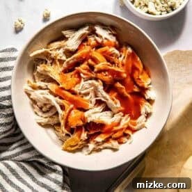 A bowl of shredded chicken topped with buffalo sauce, with a bowl of crumbled blue cheese and a striped towel nearby.