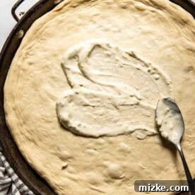 A large round pizza dough in a pan with blue cheese dressing being spread on it with a spoon.
