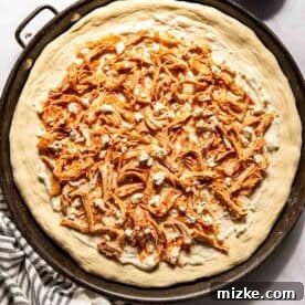 Unbaked pizza on a round tray topped with shredded buffalo chicken and crumbled cheese, with a bowl of cheese and a striped towel nearby.