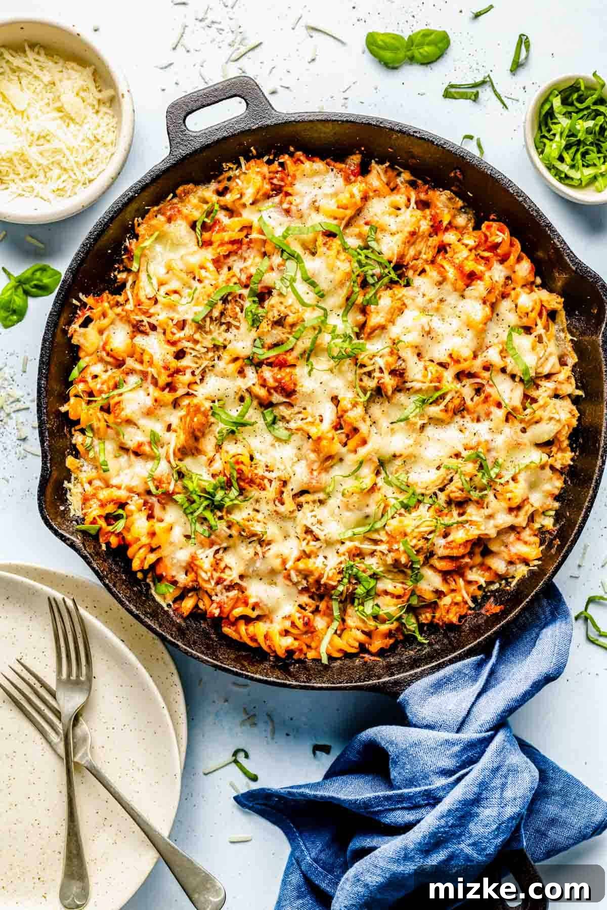 Cheesy Tuna Pasta Casserole 2 A bubbling tuna pasta bake in a cast iron skillet, garnished with fresh basil leaves, next to a bowl of grated Parmesan cheese and clean plates, ready to be served.