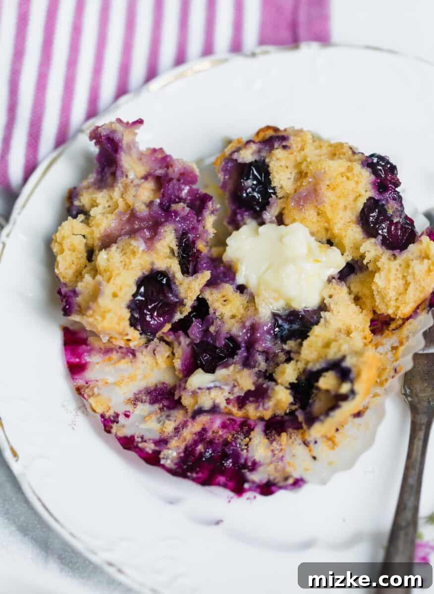 A freshly baked, warm blueberry muffin cut in half, revealing its moist crumb and juicy blueberries, with a melting pat of butter on top.