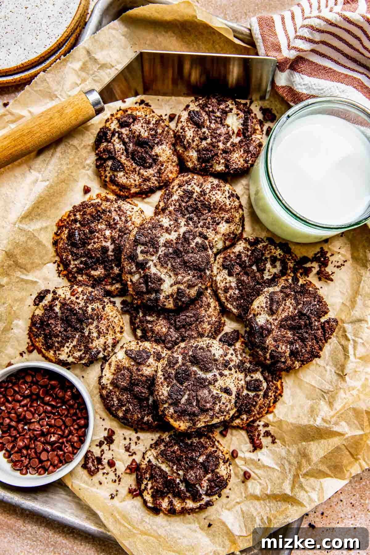 Several soft and chewy Oreo cheesecake cookies cooling on parchment paper, ready to be enjoyed, with a glass of milk and a bowl of mini chocolate chips nearby.