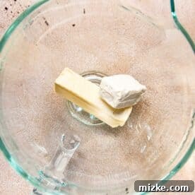 Softened cream cheese and butter being creamed together in a glass stand mixer bowl.