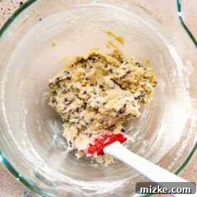 Mini chocolate chips being carefully folded into the cheesecake cookie batter with a red silicone spatula.