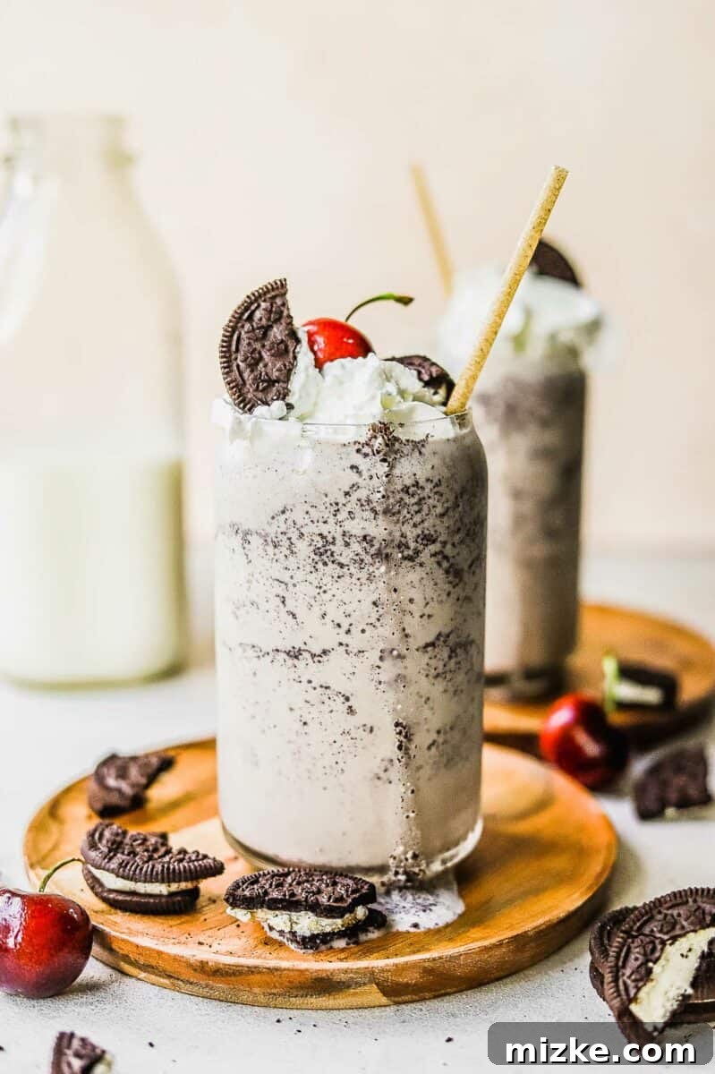 Midnight Cookie Delight 2 A large glass cup is filled with a homemade cookies and cream milkshake, topped with fluffy whipped cream, crumbled Oreo pieces, and a vibrant red cherry.