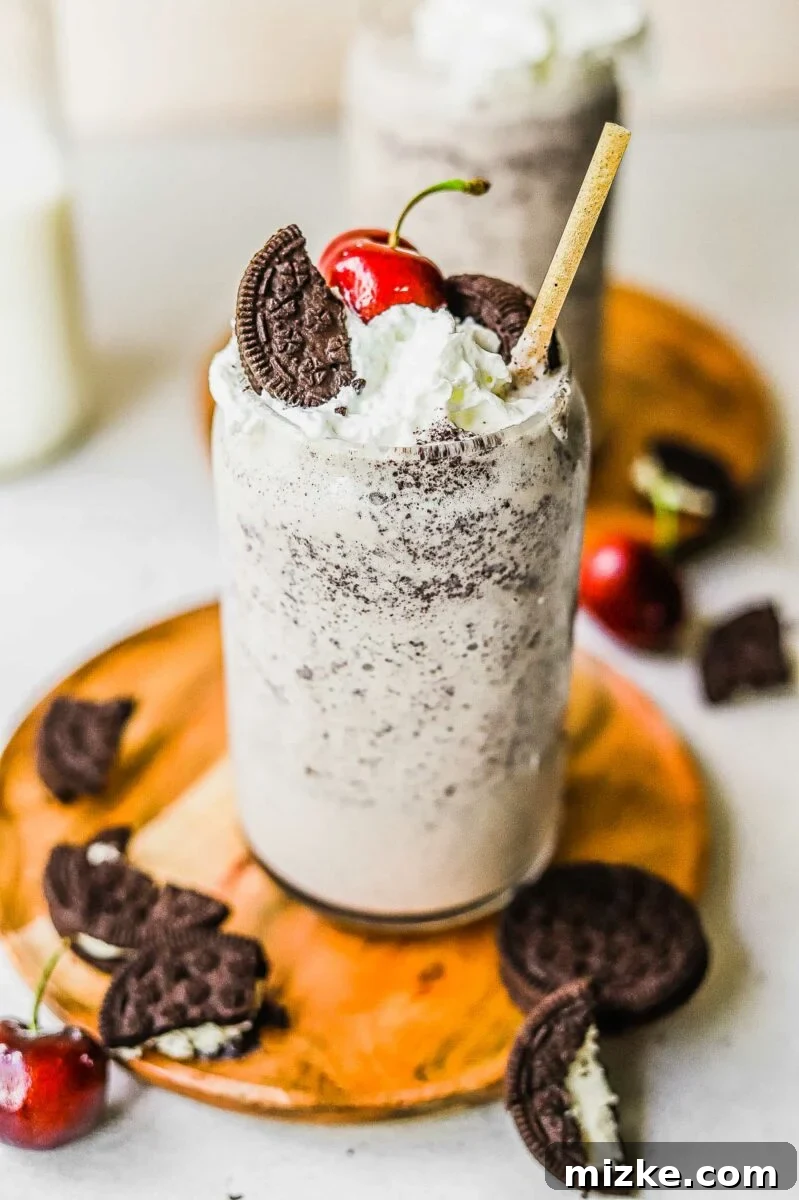 Midnight Cookie Delight 3 A clear glass of Oreo milkshake topped with a swirl of whipped cream, a single bright red cherry, and a piece of Oreo cookie, set against a blurred background.