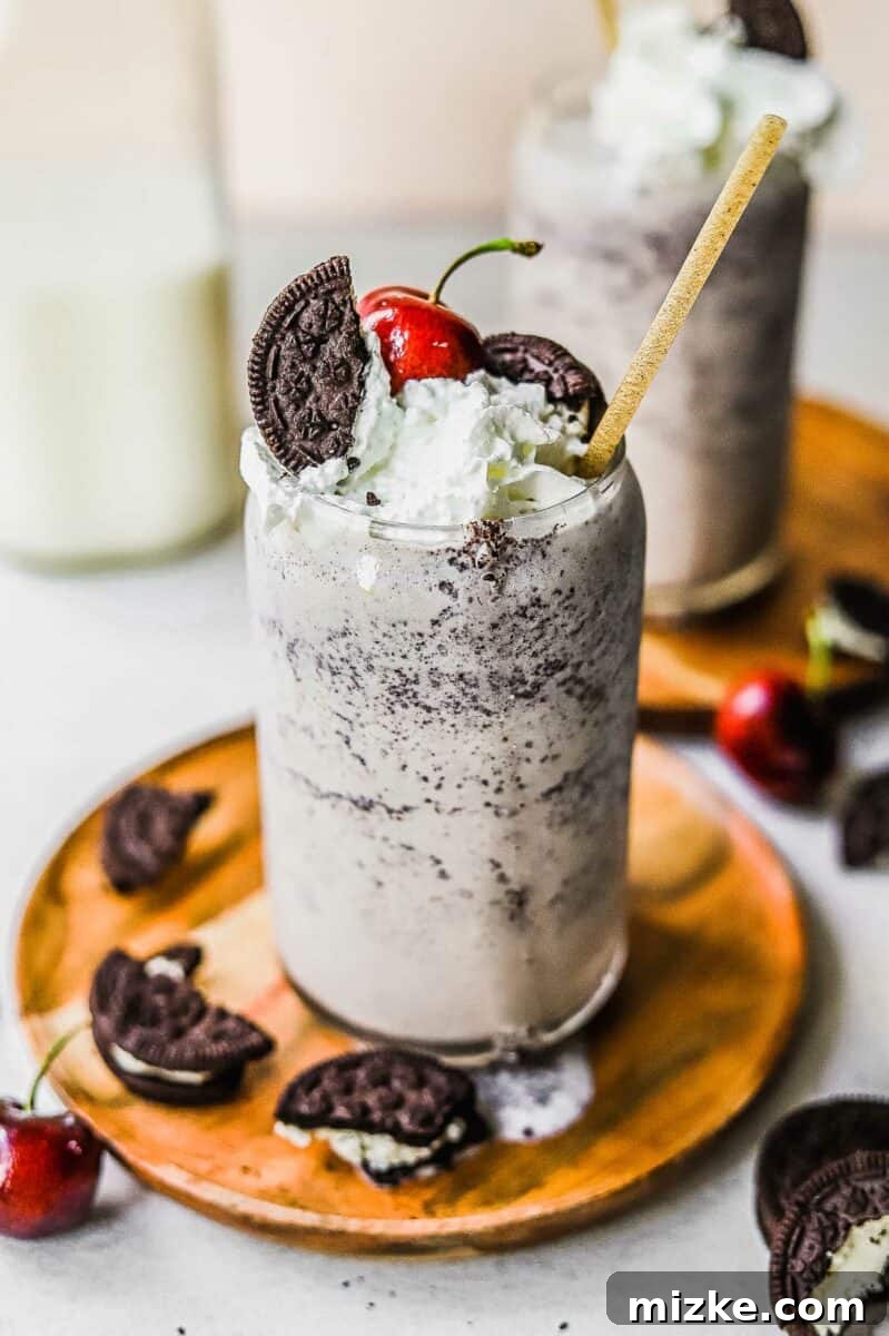 Midnight Cookie Delight 5 A blended cookies and cream milkshake is presented in a tall glass on a small wooden plate, surrounded by scattered Oreo cookie pieces.