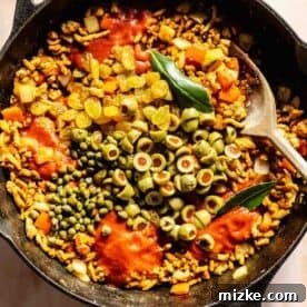 Savory Turkey Picadillo 7 Tomato sauce, golden raisins, Spanish olives, capers, and bay leaves on top of ground turkey in a cast iron skillet with a wooden spoon, illustrating the addition of wet ingredients and briny elements