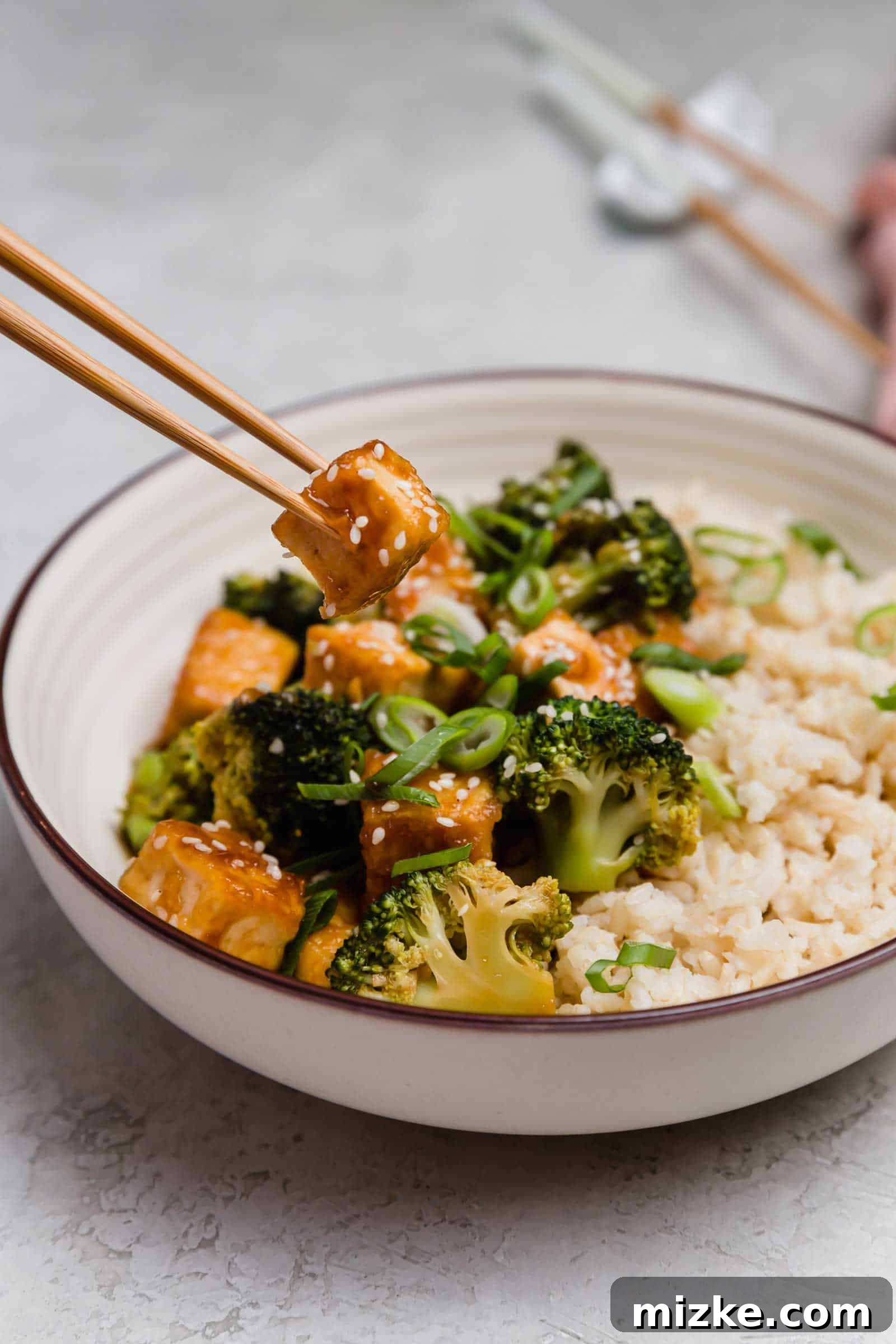 Satisfy your craving for Asian take-out with these teriyaki tofu and broccoli bowls, an easy plant-based dinner for any night of the week! Chopsticks lifting a piece of teriyaki tofu from a bowl of tofu, broccoli, and rice.