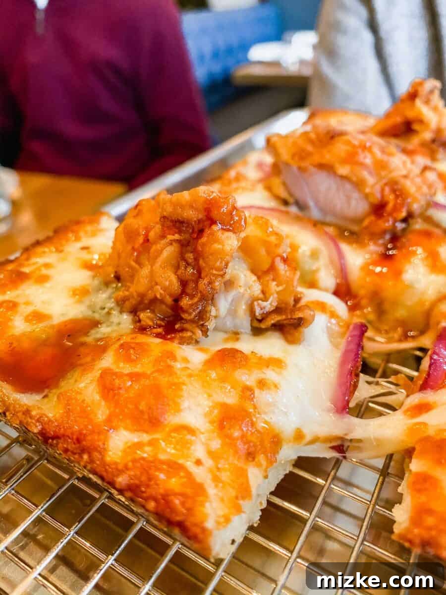 DC Date Night Done Right: Emmy Squared 8 A close-up view of a single slice of the EC Shaw pizza, highlighting the thick, crispy Detroit-style crust, melted cheese, and generous fried chicken topping.