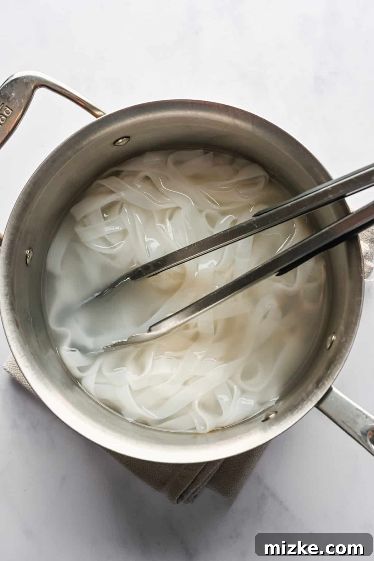 This recipe for drunken noodles with tofu is full of flavor and easy enough for a weeknight. Make your favorite Thai dish right at home! A pair of tongs resting in a pot of cooked rice noodles.