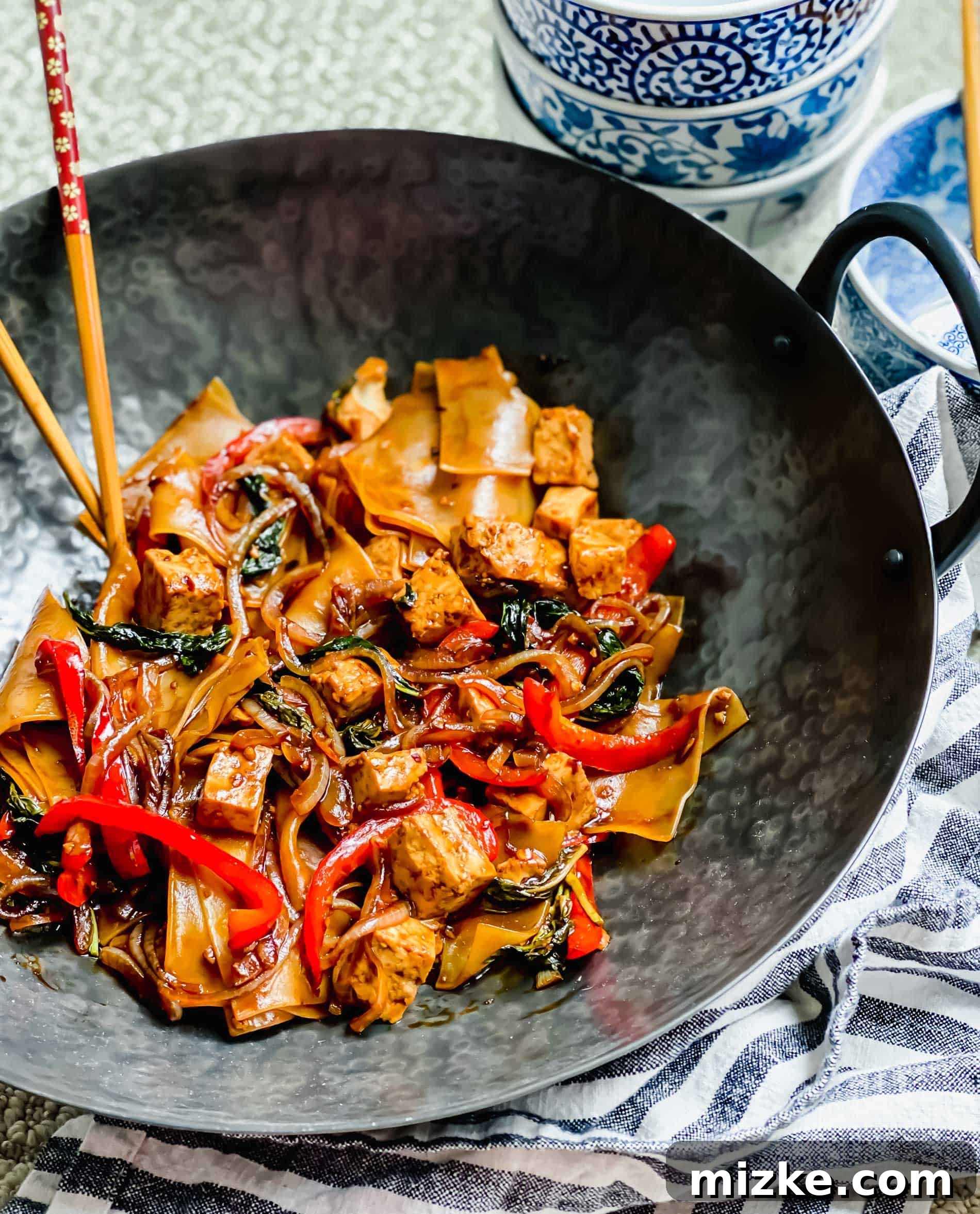 This recipe for drunken noodles with tofu is full of flavor and easy enough for a weeknight. Make your favorite Thai dish right at home! Drunken noodles in wok with with chopsticks and bowls in background.