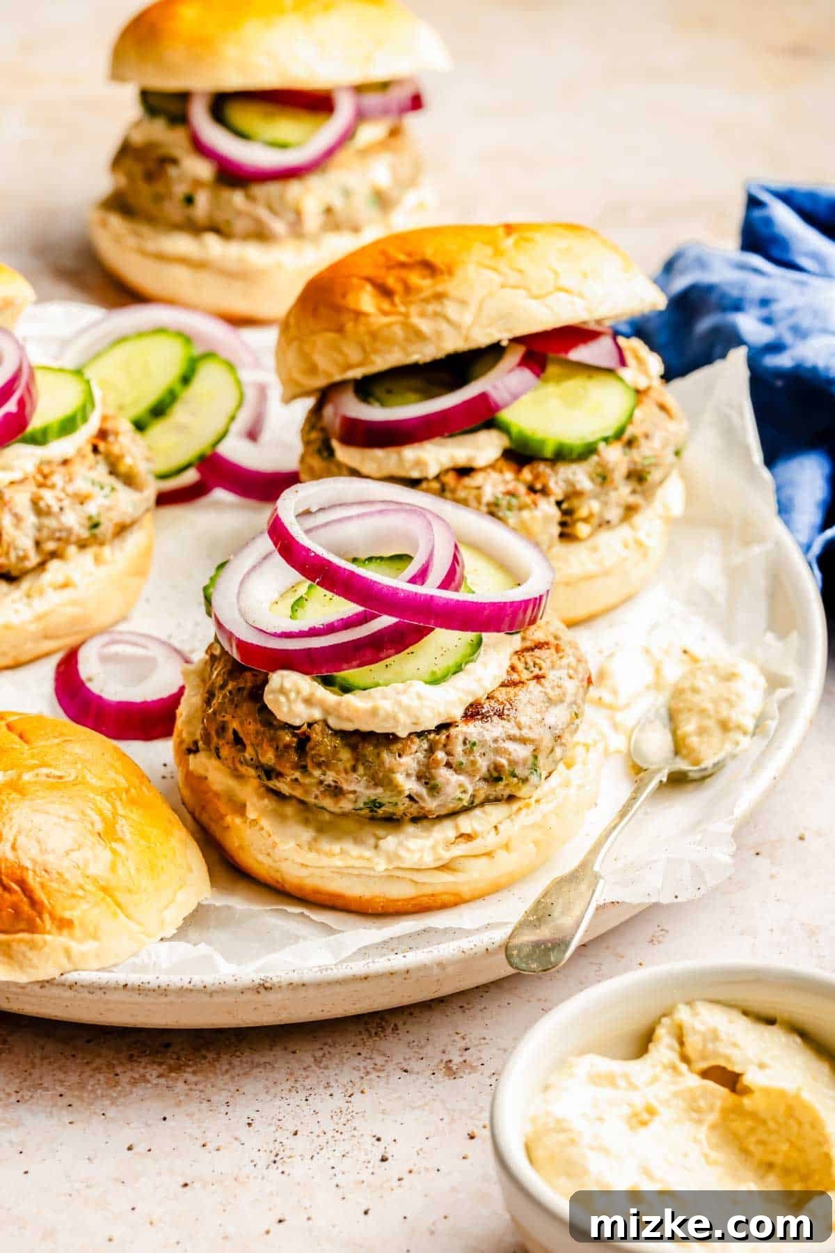 Mediterranean Turkey Spinach Feta Burgers 3 An open-faced turkey spinach feta burger showcasing its rich toppings, including creamy hummus, fresh cucumber slices, and thinly sliced red onion rings, all resting on a soft brioche bun.