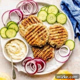 Mediterranean Turkey Spinach Feta Burgers 9 Cooked turkey spinach feta burger patties served on a large white plate, accompanied by fresh cucumbers, red onions, and a bowl of creamy hummus, ready for assembly.