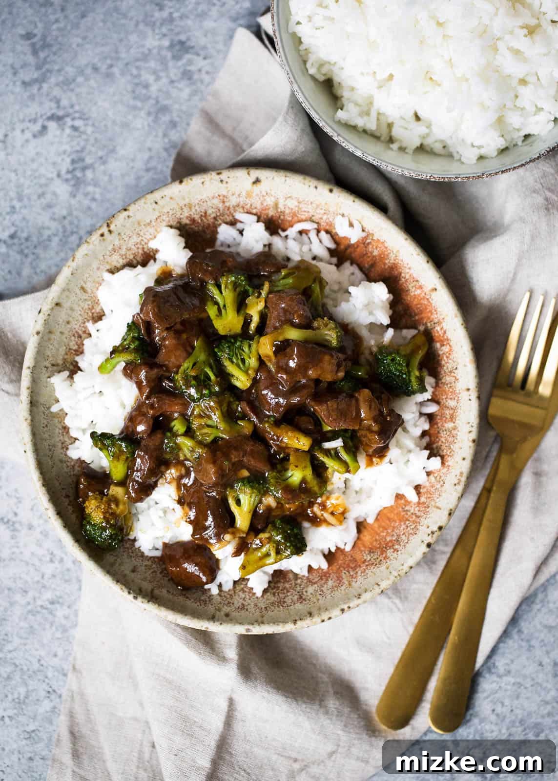 Easy Slow Cooker Beef and Broccoli 3 An inviting overhead shot of slow cooker beef and broccoli generously spooned over a bed of white rice in a tan speckled bowl with a rust-colored interior. The bowl rests elegantly on a beige linen towel, accompanied by a gleaming gold fork and knife, and another bowl of pristine white rice visible in the upper right background, completing a harmonious dining scene.
