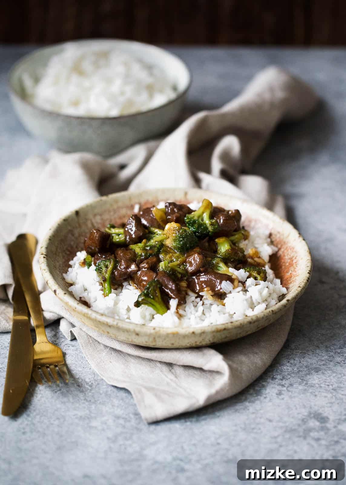 Easy Slow Cooker Beef and Broccoli 4 A beautifully presented serving of slow cooker beef and broccoli alongside white rice, nestled in a tan speckled bowl with a rust-colored rim. The bowl sits on a beige linen towel, flanked by a shining gold fork and knife, with an additional bowl of fluffy white rice visible in the background, completing a rustic and inviting culinary scene.