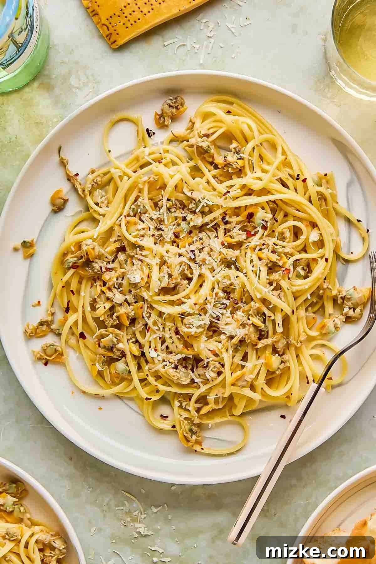 White Wine Clam Linguine 2 A beautifully plated dish of linguine with clams, generously garnished with freshly grated Parmesan cheese and a sprinkle of vibrant red pepper flakes. A fork rests beside the plate, ready to be enjoyed.