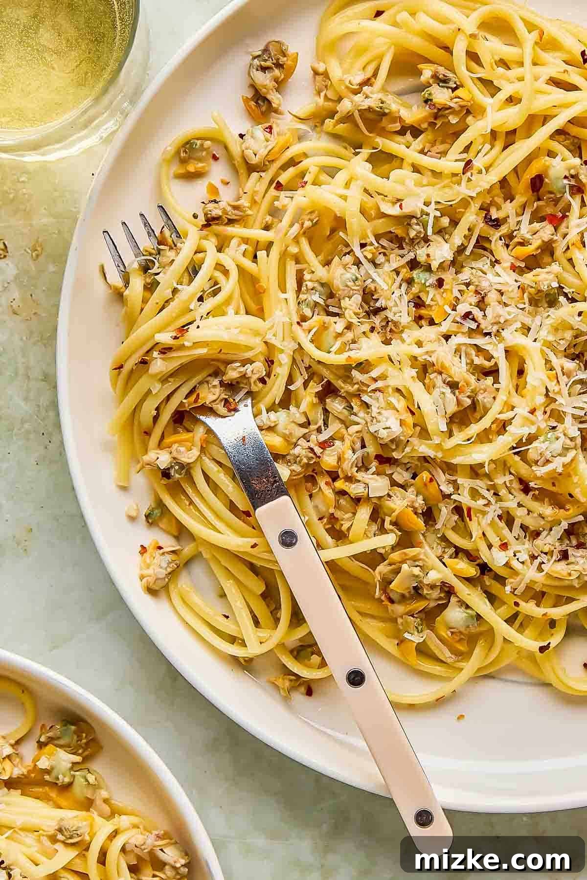 White Wine Clam Linguine 4 A vibrant plate of linguine with clams, beautifully adorned with grated cheese and red pepper flakes. A fork rests delicately on the pasta, while a glass of crisp white wine is partially visible in the background, hinting at a perfect pairing.