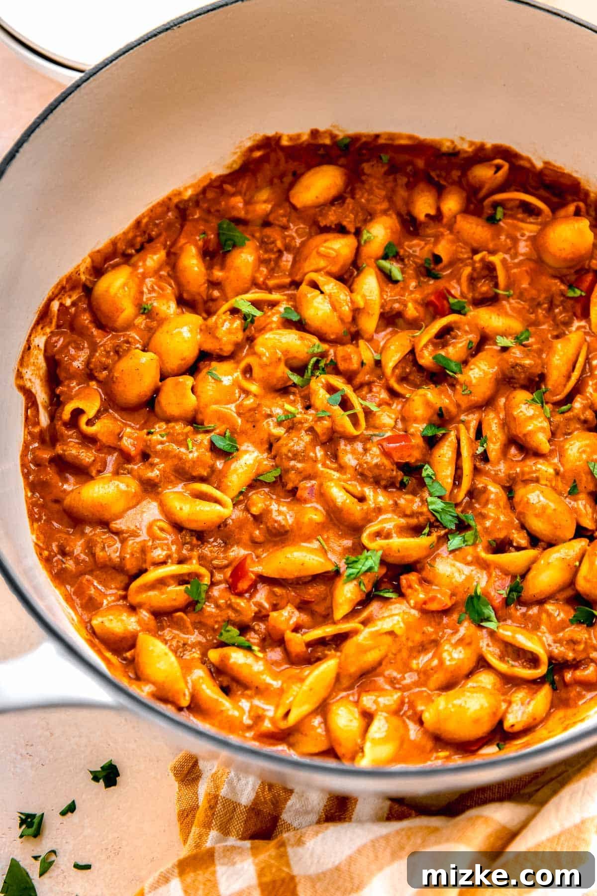 Ground beef with pasta shells in a cast iron skillet next to a yellow checkered linen towel with fresh parsley garnished on top, ready to be served.