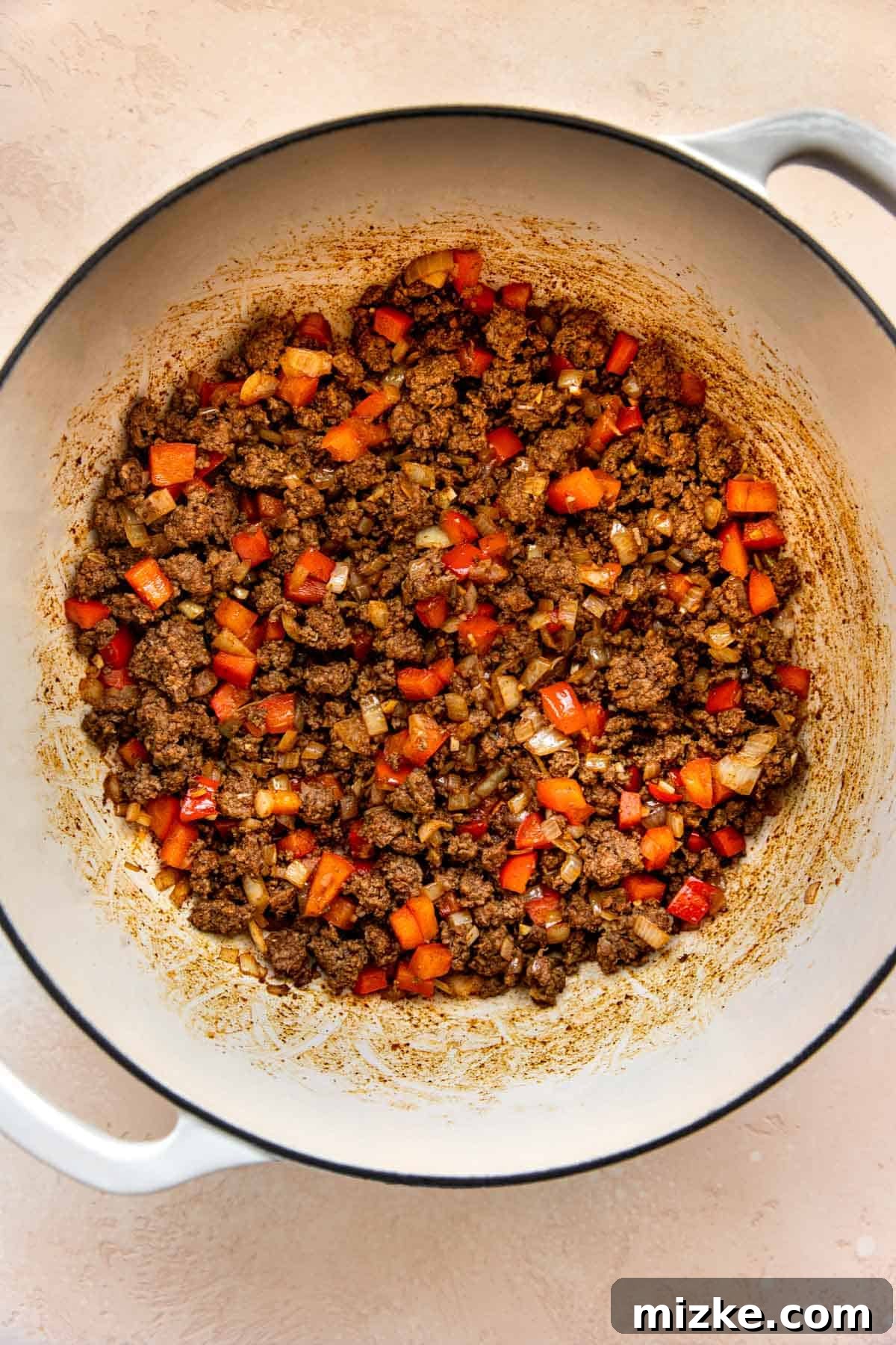 Browned ground beef mixed with red bell peppers and onions in a cast iron skillet.
