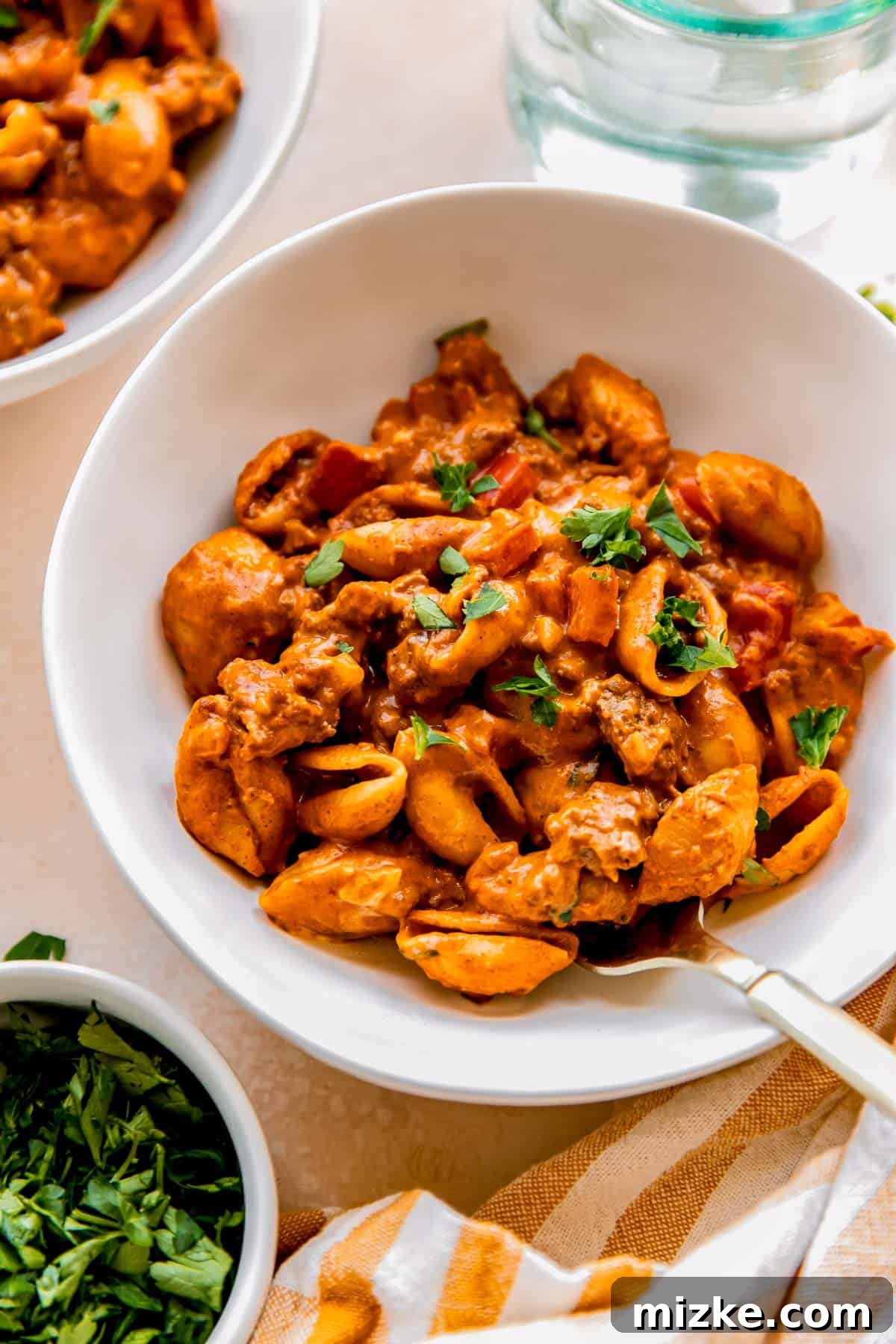 A white bowl filled with creamy ground beef and pasta shells, garnished with fresh parsley and a metal fork.