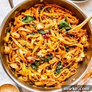 Sun-dried tomato chicken pasta in a stainless steel skillet with a wooden spoon next to a yellow linen towel