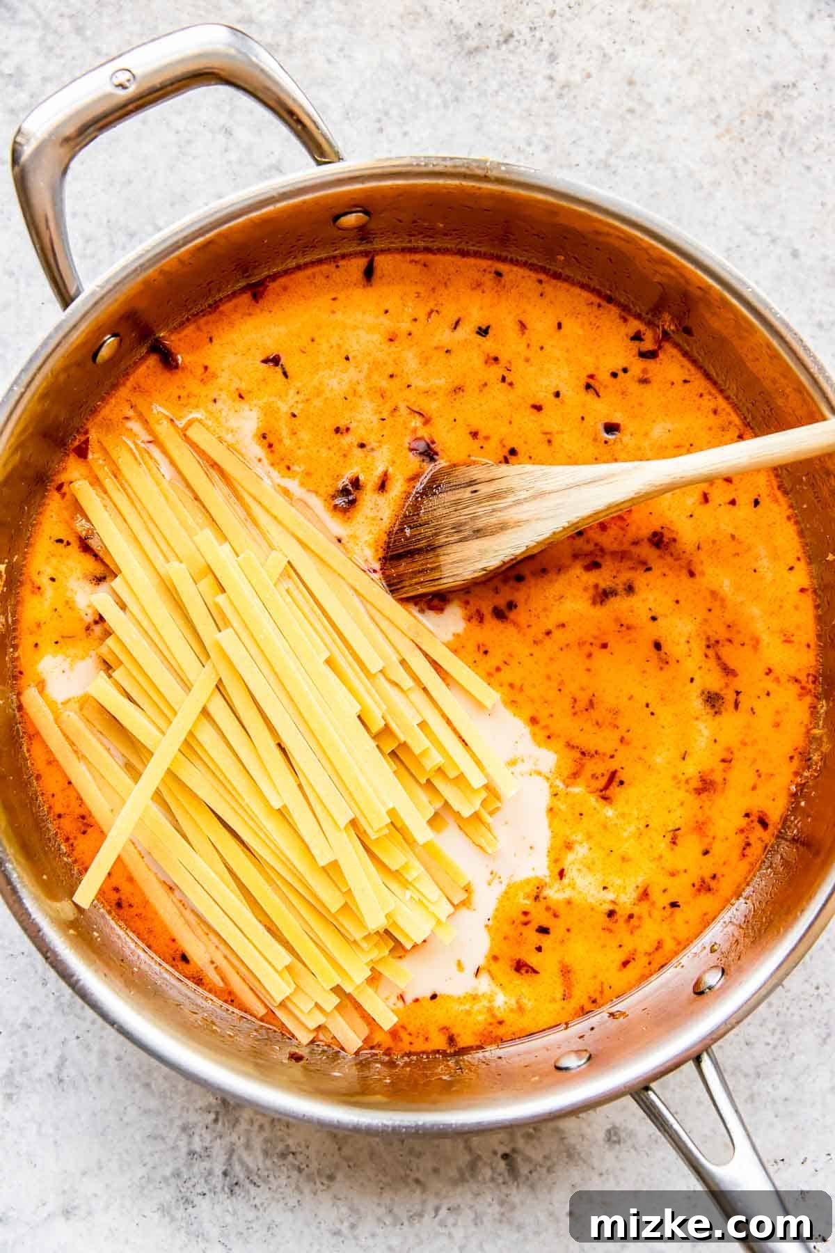 Sun-Kissed Skillet Chicken Pasta 9 Dry linguine pasta carefully added to the stainless steel skillet, submerging into the creamy liquid with a wooden spoon nearby