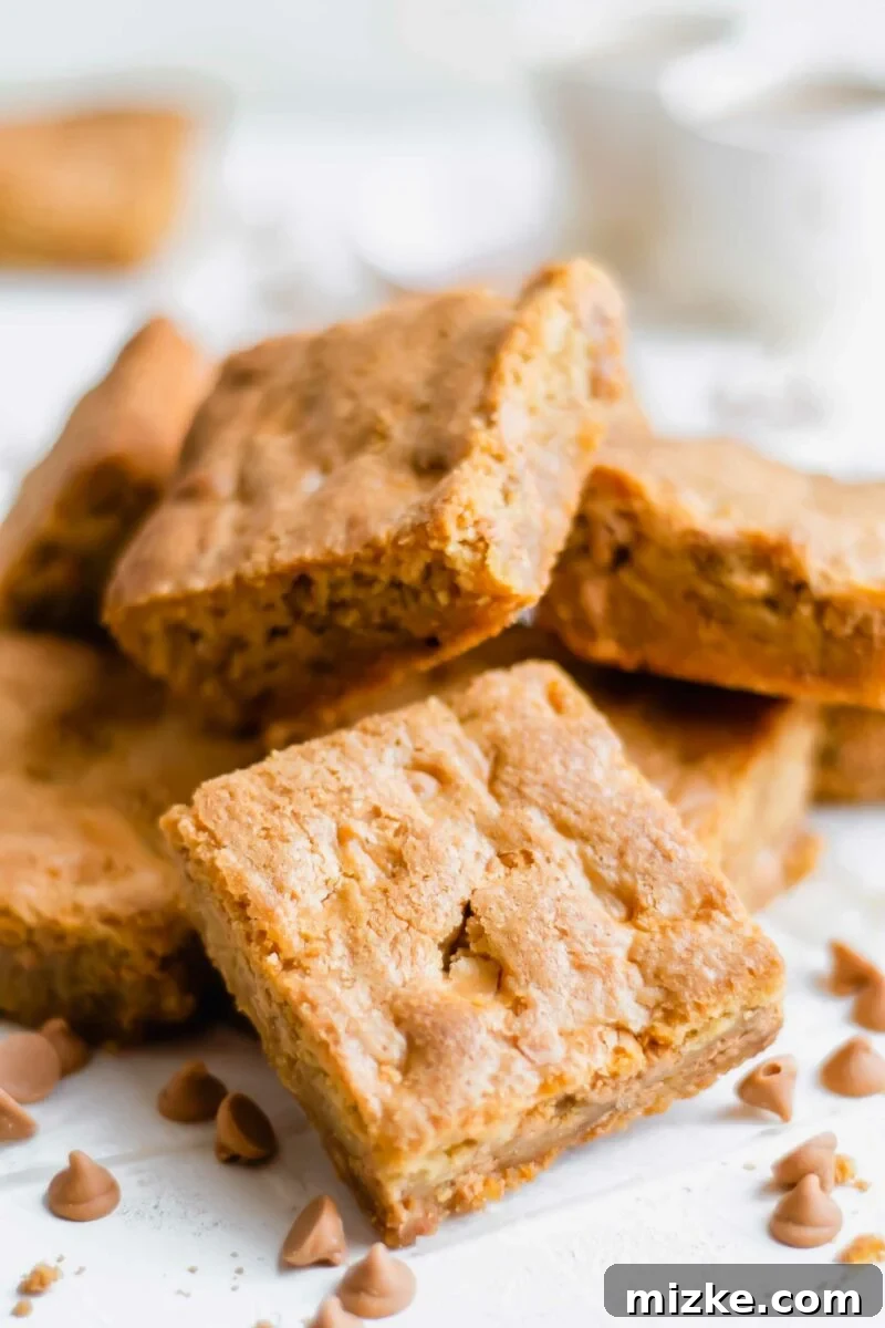 Golden Chewy Blondies 6 A stack of golden butterscotch blondies on a white surface, surrounded by loose butterscotch chips, emphasizing the key flavor of the dessert.
