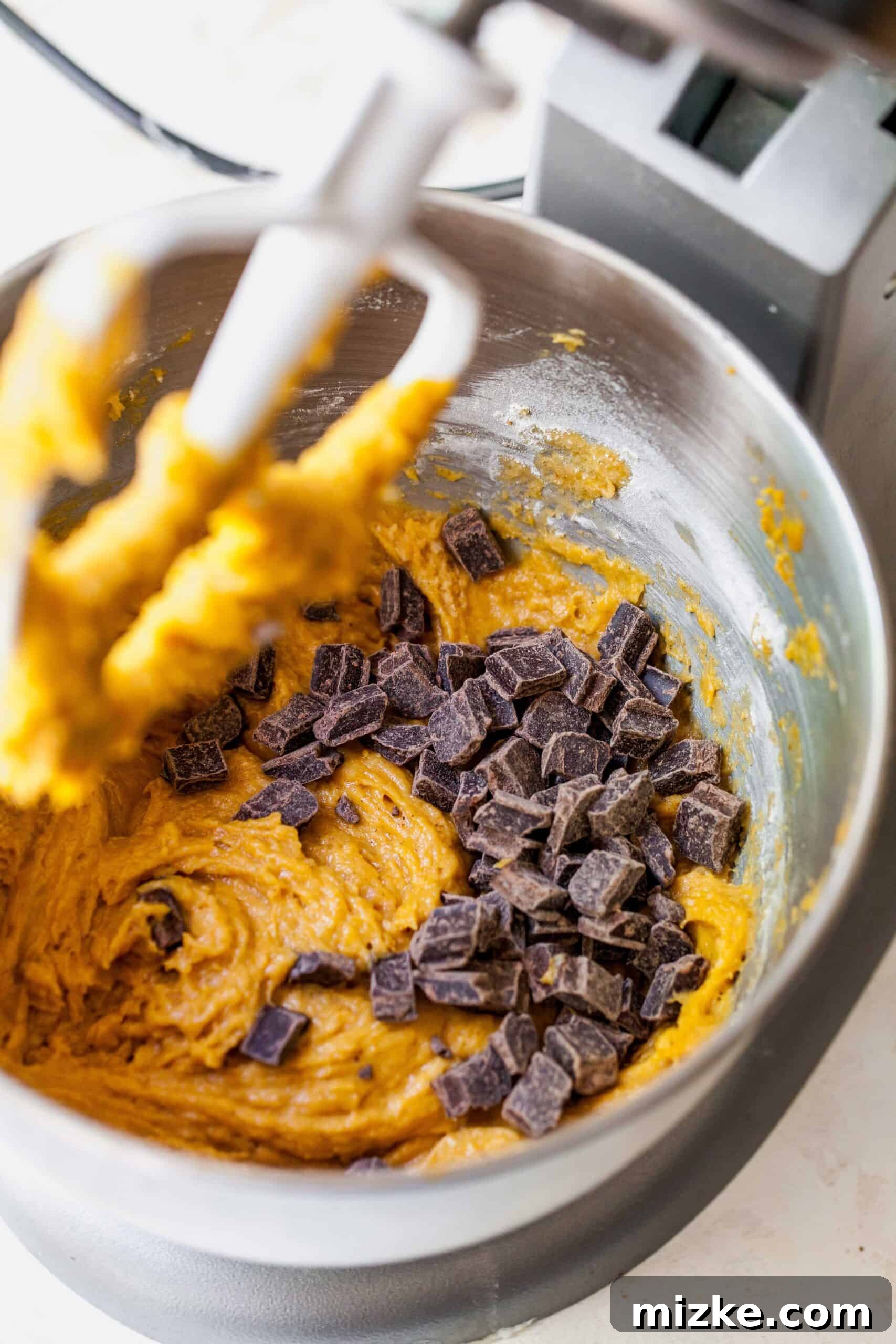 Close-up of luscious semi-sweet chocolate chunks being carefully added to a vibrant pumpkin muffin batter in a stand mixer, ready to be folded in.