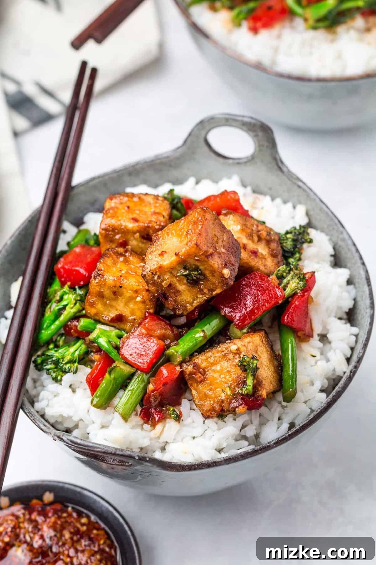 Crispy Tofu Stir-Fry 13 A beautifully presented bowl of spicy chili tofu stir fry and white rice is artfully placed beside a small condiment bowl filled with rich chili crunch sauce on a pristine white countertop, ready to be enjoyed.